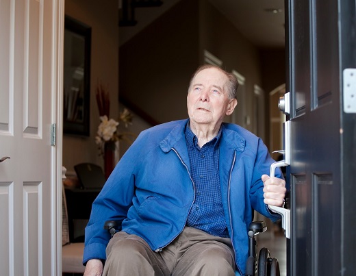 Old man sitting on the wheel chair and holding door's handle and looking outside the home